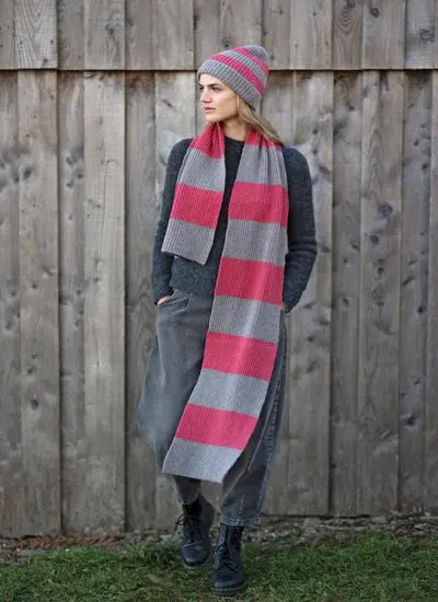 blonde woman standing against wooden fence wearing a large grey and pink striped knit scarf